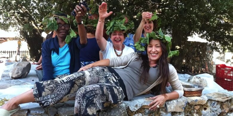 A group of people smiling and waving, wearing grapevine wreaths on their heads, with one person playfully lying across a stone surface in front.