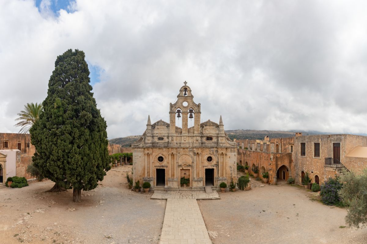 Arkadi Monastery in Crete, featuring a historic stone facade with arched doorways and bell towers. The courtyard is surrounded by old buildings, with a large tree on the left and a cloudy sky overhead.