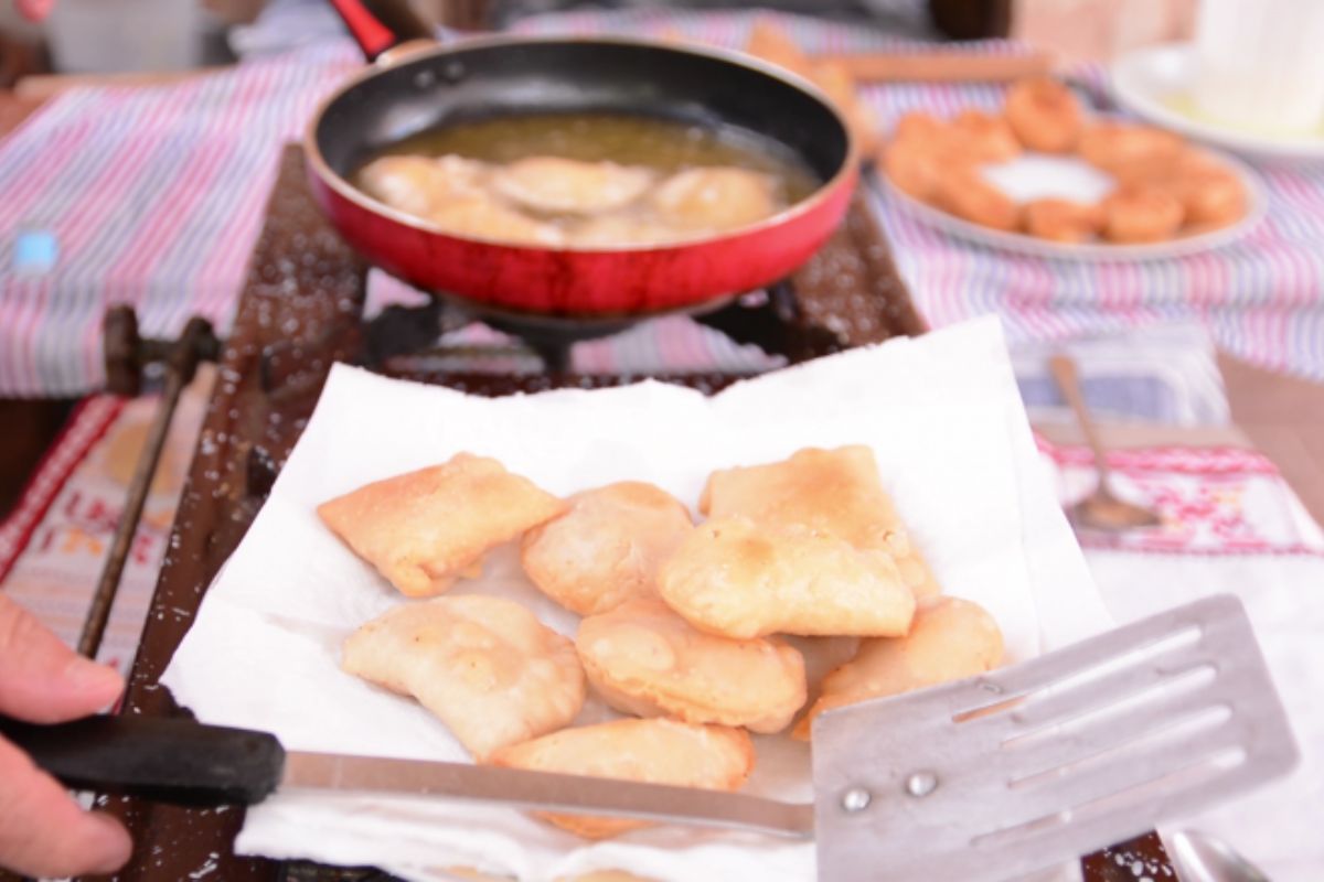 A Crete cooking class where golden, freshly fried pastries are being prepared. A spatula holds the pastries on a paper towel, while a pan of hot oil and more treats are visible in the background.