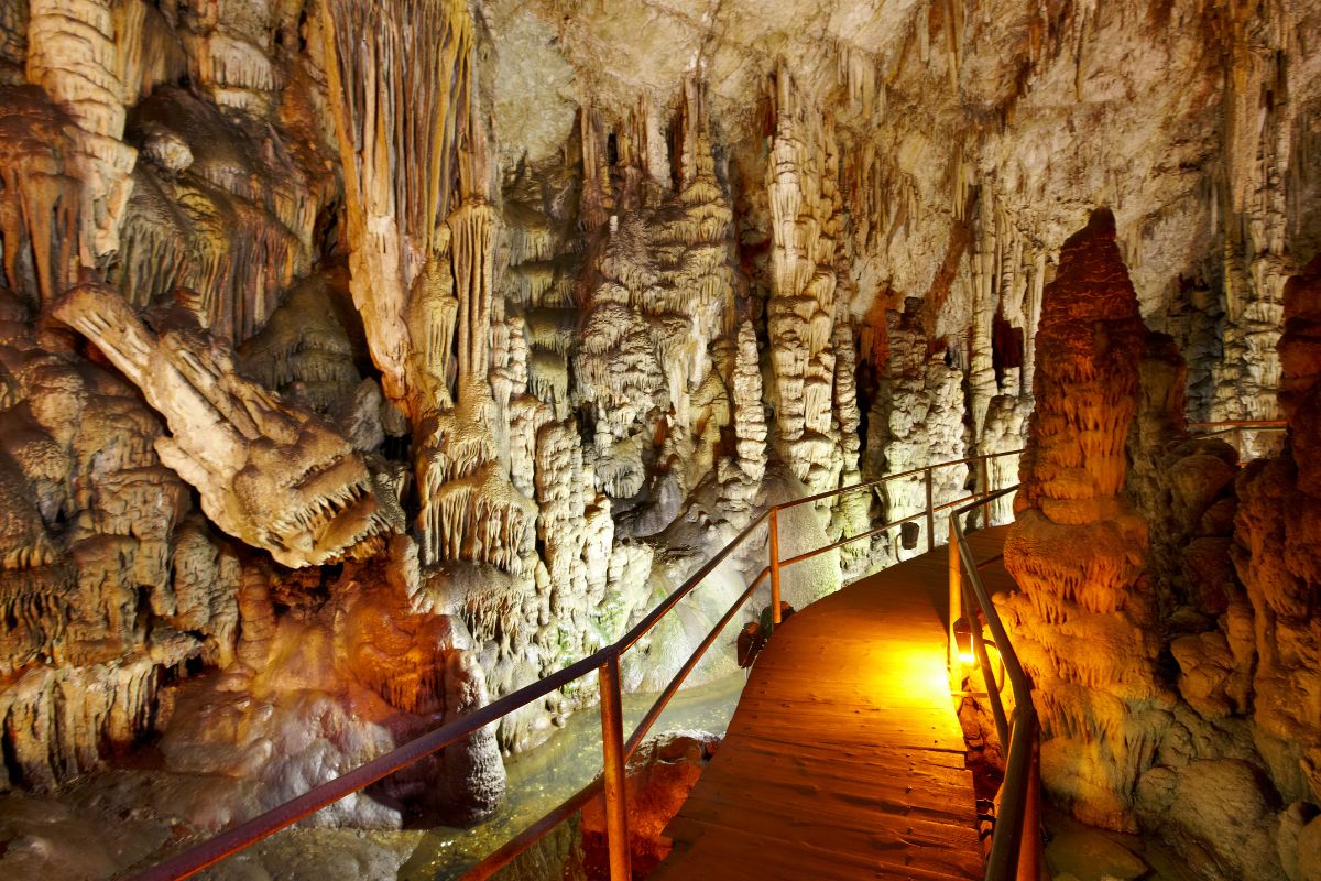 Dikteon Cave, a limestone cavern in Crete, featuring stalactites and stalagmites. A wooden walkway with railings winds through the illuminated rock formations.