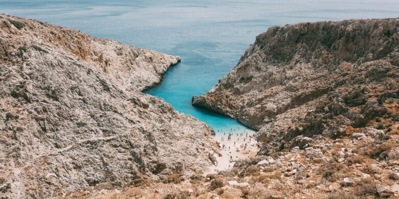 Seitan Limani Beach in Crete, a small sandy cove surrounded by steep rocky cliffs. The turquoise water meets the shore where people are swimming and sunbathing.