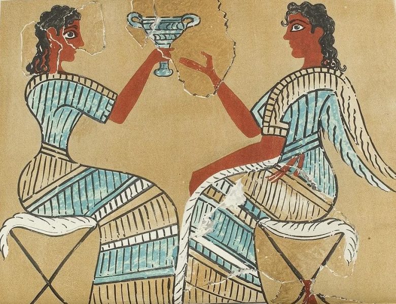 The image is a fragment of the Campstool fresco from Knossos, depicting two Minoan women. The women, with intricately styled hair and wearing traditional Minoan attire characterized by fitted bodices and flounced skirts, are seated on stools with crossed legs. One woman is raising a cup, while the other gestures with an open hand, possibly in conversation. The fresco is rendered in earthy tones with details in blue and red, reflecting the artistry of the ancient Minoan civilization. Some parts of the fresco are damaged, revealing the historical wear of this archaeological find.