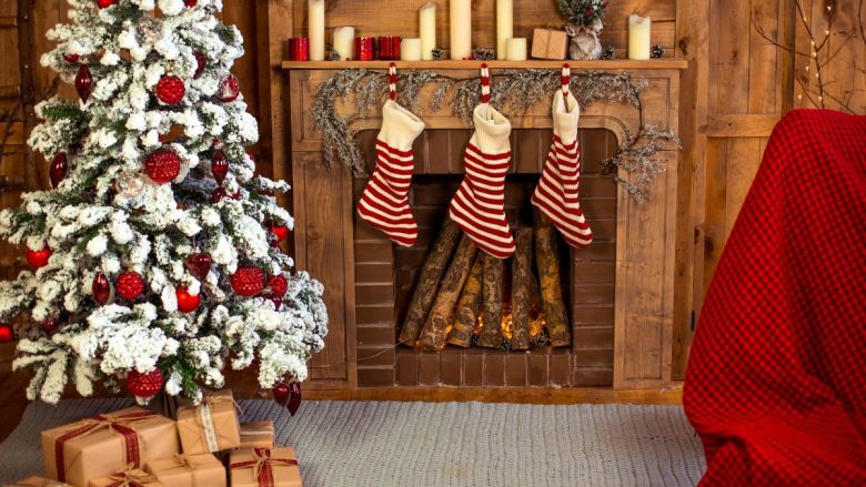 The image depicts a cozy Christmas setting with a beautifully adorned Christmas tree dusted with snow-like decorations and bright red ornaments. Three striped Christmas stockings hang on the mantle of a rustic fireplace, where logs are ablaze, adding warmth to the scene. Wrapped presents sit at the base of the tree, and a comfy chair with a red checkered blanket suggests a perfect spot for relaxing in this festive room.