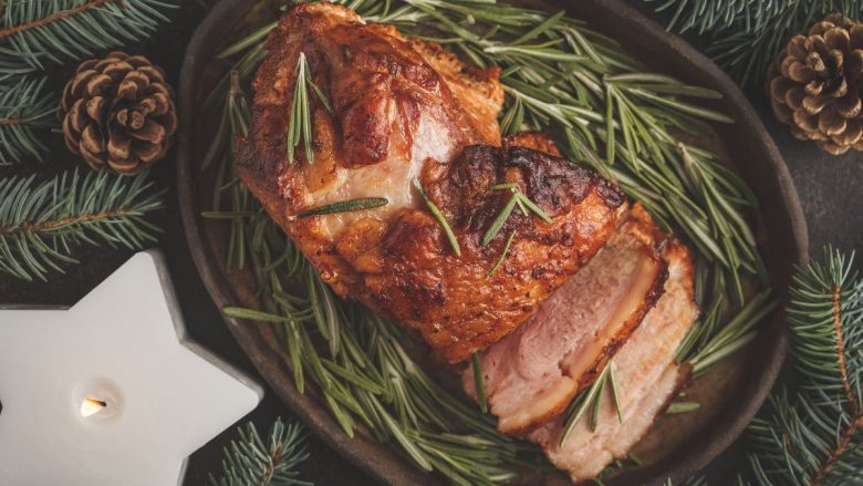 The image displays a roasted pork loin, garnished with sprigs of fresh rosemary, presented in a rustic cooking pan. It's surrounded by festive decorations including pine branches and a pine cone, alongside a white star-shaped candle, evoking the cozy ambiance of a Christmas meal. This setting is indicative of a traditional Cretan Christmas feast.