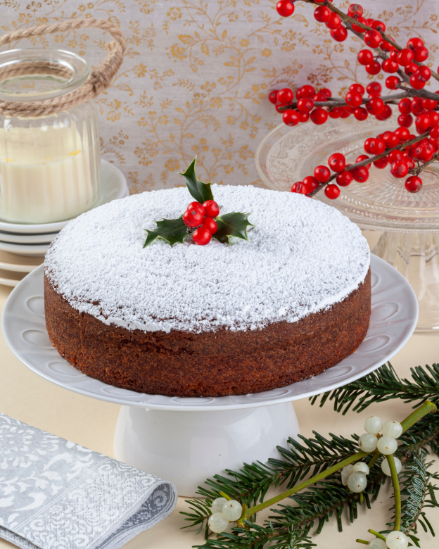 The image shows a traditional Vasilopita, which is a Greek New Year's cake, dusted with powdered sugar and topped with a sprig of holly. The cake is presented on a white pedestal plate, set against a festive backdrop with a patterned cloth, a candle, and red berries. This confection is part of the New Year celebrations in Crete, symbolizing good fortune for the year ahead.
