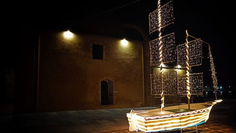 The image shows a traditional Cretan "karavaki", a model boat adorned with lights, set against a night backdrop. The boat is decorated with white lights outlining its form and with panels that could be sails, illuminated by a string of lights. This display is set up on a pavement, in front of a building with a plain facade and lit windows, capturing the essence of the festive season in Crete.