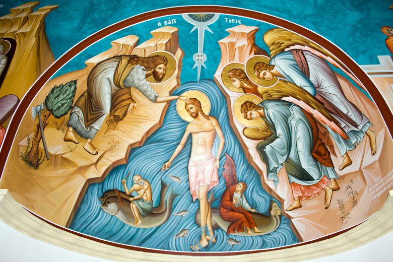 This is an image of a colorful mural depicting the baptism of Jesus Christ. In the center, Jesus stands in a river with a halo around his head, and John the Baptist is shown pouring water over Jesus' head with one hand while holding a cross staff in the other. Above Jesus, a dove, symbolizing the Holy Spirit, descends with rays of light. Surrounding them are four angels, each holding a garment, and on the left side, an elder man with an axe stands by a tree. The river is animated with fish, and a man is depicted riding a fish at the bottom left. The artwork is done in a traditional Orthodox iconographic style with vibrant colors and gold accents, and the scene is set within a semi-circular arch.