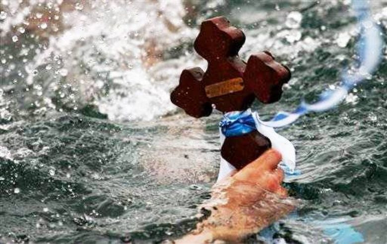 The image captures the dynamic moment of a traditional Greek Epiphany celebration with a person's arm reaching out of the water to grasp a wooden cross. The cross is adorned with a blue and white ribbon, reflecting the colors of the Greek flag. Splashes of water surround the action, indicating the vigor of the retrieval ritual during the Blessing of the Water ceremony, which is part of the Epiphany festivities in Greece.