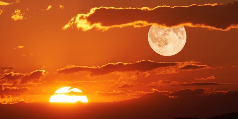 A vivid image depicting a dramatic sunset with the sun touching the horizon beneath a large, detailed moon in the orange-hued sky. Clouds are scattered across the sky, outlined radiantly by the sun's glow. The scene captures the dual presence of both sun and moon, symbolizing the passing of time, likely resonating with themes of the Minoan Sacred Year.