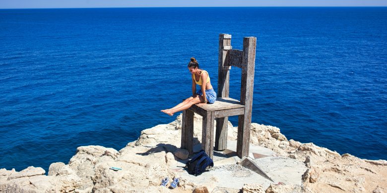 Person sitting on a large wooden chair on a rocky shore overlooking the sea.