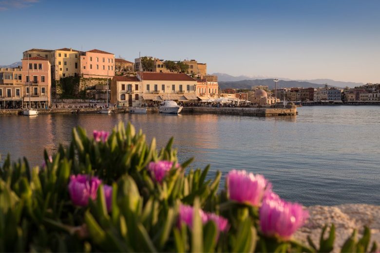 A scenic view of Chania's waterfront with buildings and a calm harbor, framed by flowering plants in the foreground.