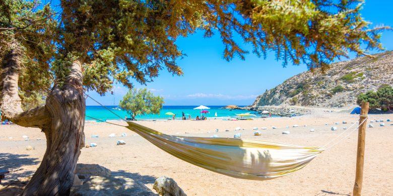 Beach view with a hammock tied between trees, umbrellas, and people enjoying the seaside.