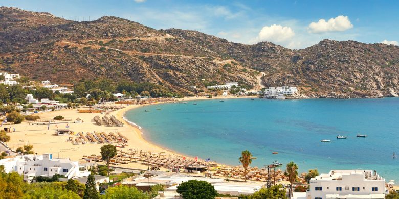 A coastal view showing a beach with loungers, clear blue waters, and surrounding hills.