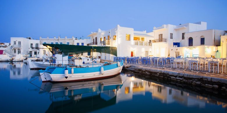 Evening view of a calm marina with moored boats, reflecting on the water, adjacent to white buildings with outdoor seating.