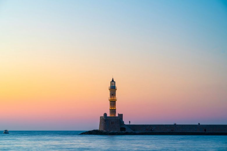 A lighthouse stands against a sunset sky with soft hues of orange and blue over a calm sea.
