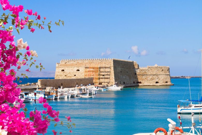 A view of a historic stone fortress by a harbor with boats and a clear blue sky, framed by pink flowers in the foreground.