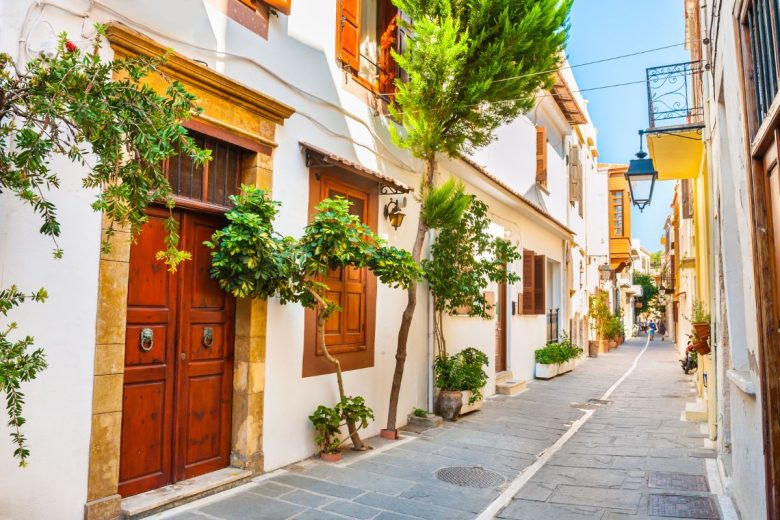 A narrow street lined with traditional houses and greenery in a sunny Mediterranean setting.
