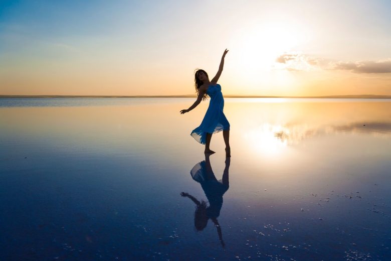 A silhouette of a person dancing by a calm water body during sunset, reflecting on the surface.