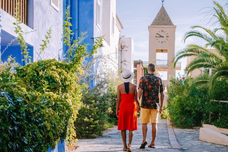A couple holding hands while walking down a quaint street lined with vibrant plants, with a clock tower in the background.