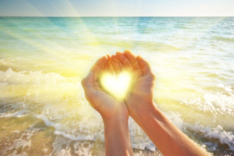 Hands forming a heart shape against a sunny beach backdrop.