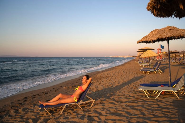 A person relaxing on a lounge chair on a sandy beach at sunset, with straw umbrellas and the ocean in the background.