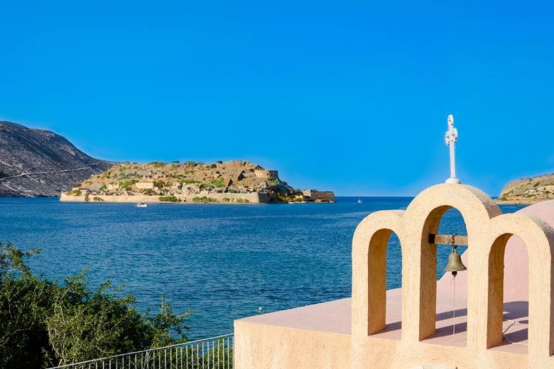 A view of the sea with a small island in the distance, seen from behind a pale arched structure with a bell and a cross.