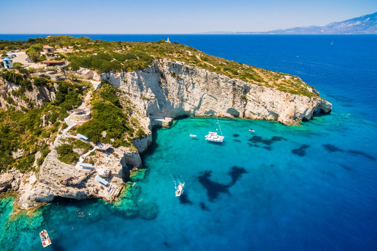 A coastal cliffside with lush greenery, crystal-clear blue water, and several boats anchored near the shore, under a clear sky.