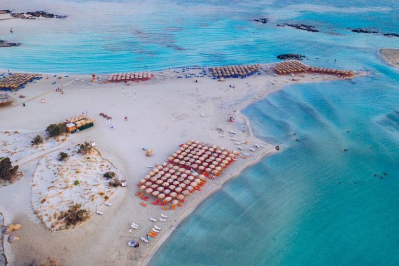 An aerial view of Elafonissi Beach in Crete, showing a sandy shore with organized sunbeds and umbrellas, surrounded by clear, turquoise waters. 
