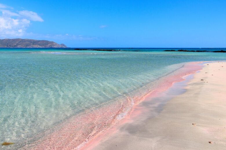 A view of Elafonisi Beach in Crete, showcasing its clear turquoise waters and a stretch of shoreline with pinkish sand. In the background, there are distant hills under a blue sky.