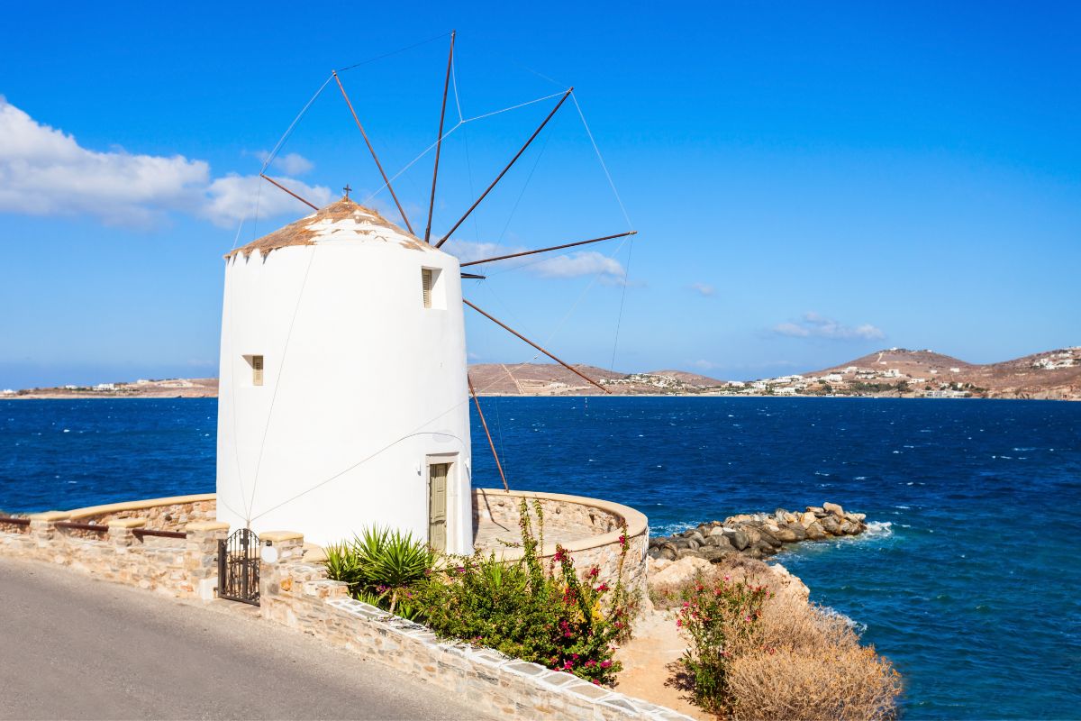 A traditional white windmill on Paros Island, Greece, overlooking the deep blue Aegean Sea with a clear sky and distant hills in the background.