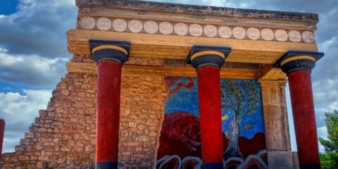 A section of the Knossos Palace featuring red columns, a stone wall, and a colorful fresco with a bull and a tree, set against a partly cloudy sky.