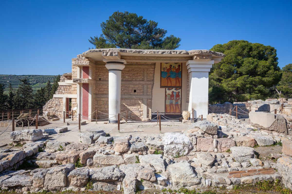 A section of Knossos Palace featuring white columns, stone walls, and a fresco depicting figures in blue and red, surrounded by ruins and greenery under a clear blue sky.