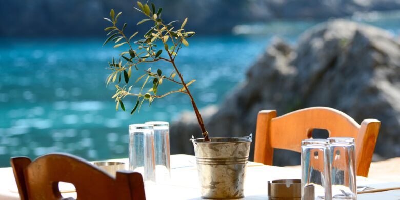 Table at a seaside Cretan taverna with an olive branch centerpiece and glasses, overlooking turquoise waters.