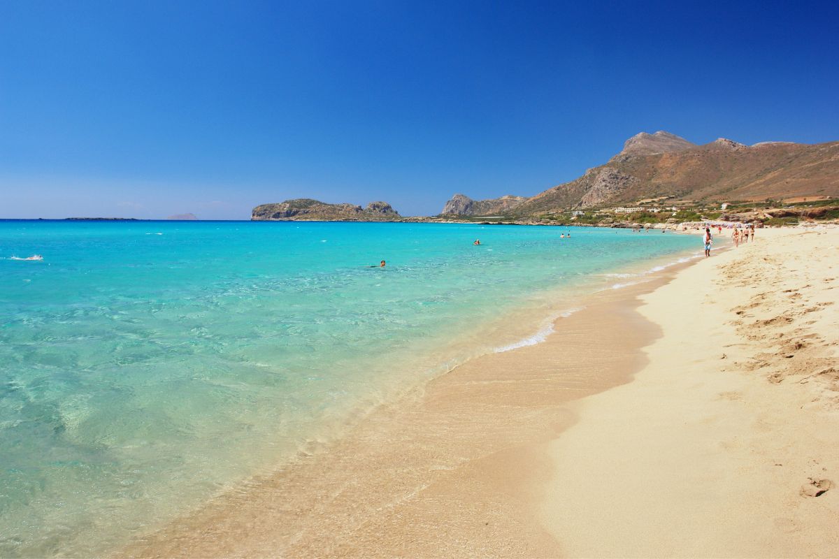 Falassarna Beach in Chania, Crete with clear turquoise water, soft sand, and people swimming and walking along the shore