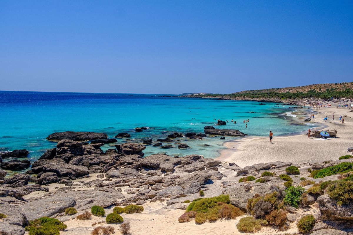 Kedrodasos Beach in Chania, Crete with turquoise waters, rocky coastline, and people swimming and walking along the shore