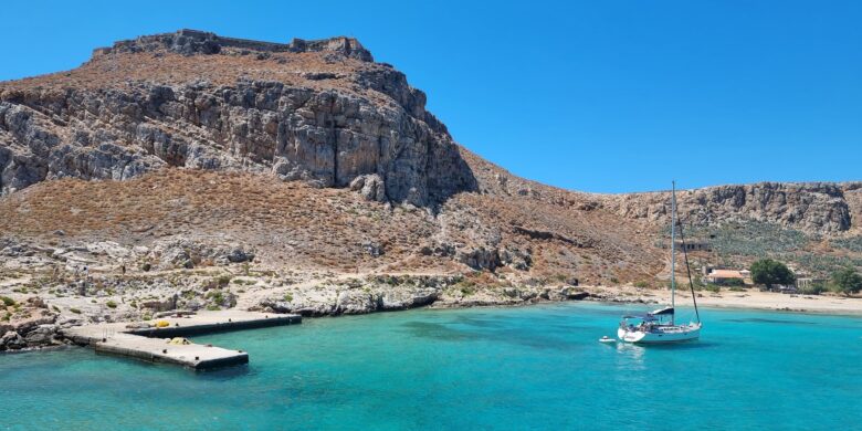 Sailboat anchored near a rocky coastline in Crete in September with clear blue waters and a hillside fortress in the background.