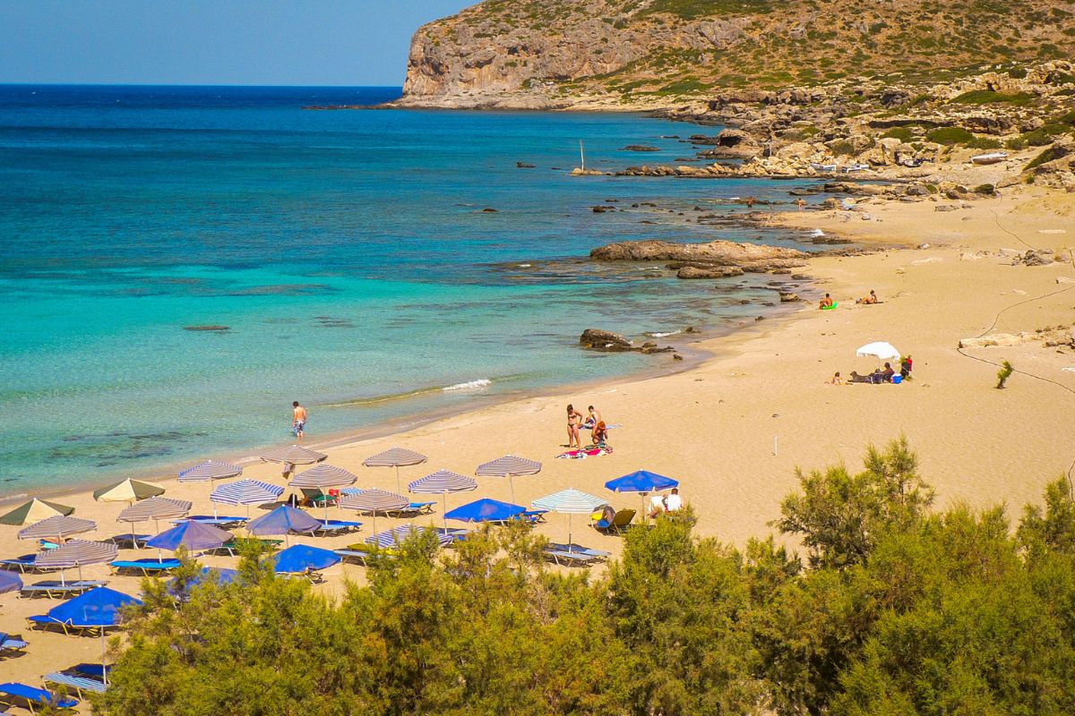 Falasarna Beach in Crete with clear turquoise waters, sunbeds, umbrellas, and visitors enjoying the sandy shore.