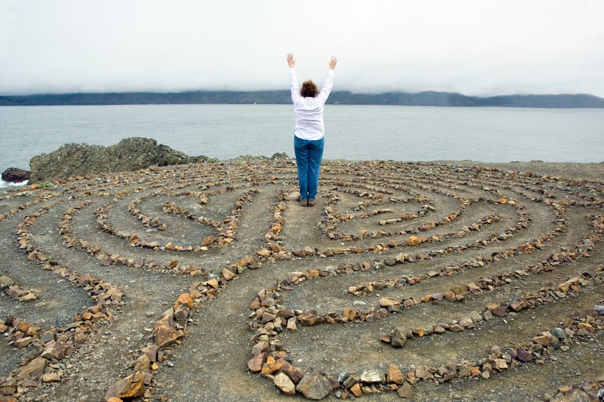 Person standing in a stone labyrinth by the sea, symbolizing Divine Feminine Goddess initiation, cycles, and inner transformation.