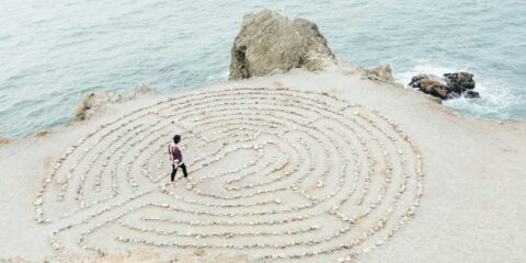 Person walking a seaside stone labyrinth, representing Divine Feminine Goddess symbolism of inner journey, cycles, and transformation.