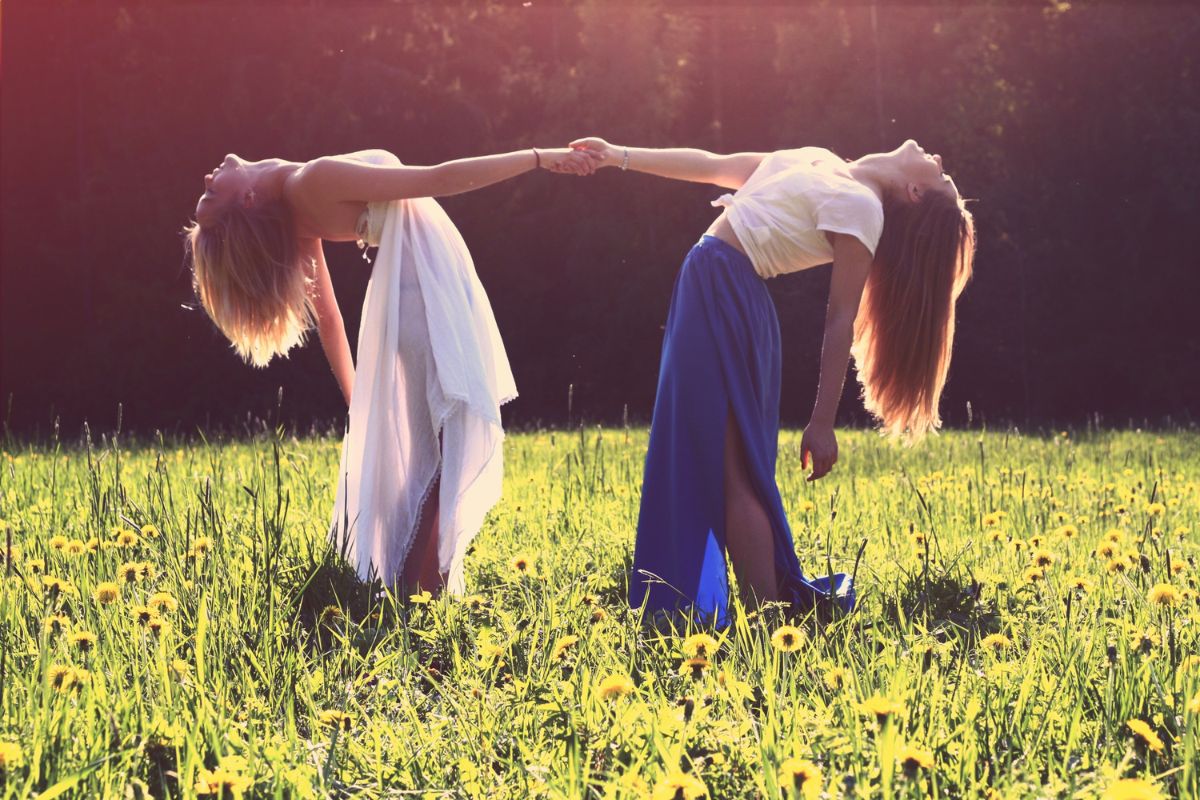 Two women in flowing dresses hold hands in a sunlit meadow, evoking Divine Feminine Goddess energy and connection with nature.