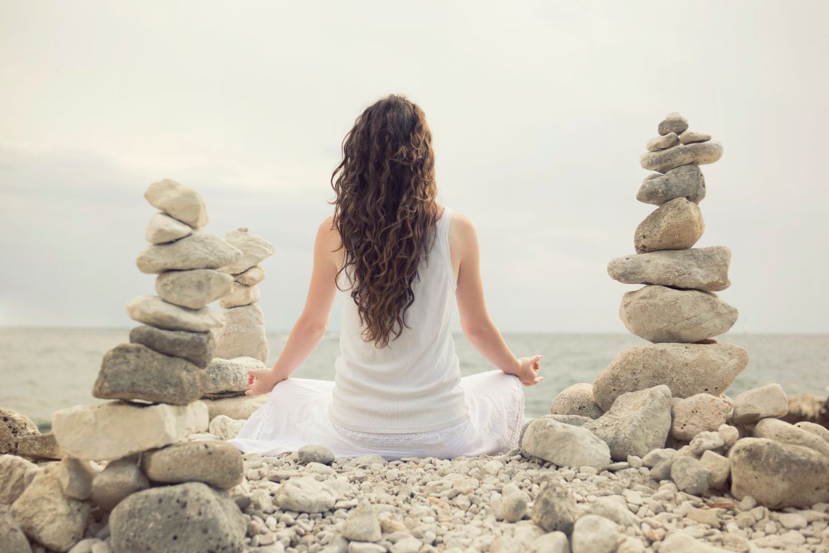 Woman meditating by the sea between stacked stones, symbolizing healing the feminine energy and inner balance.