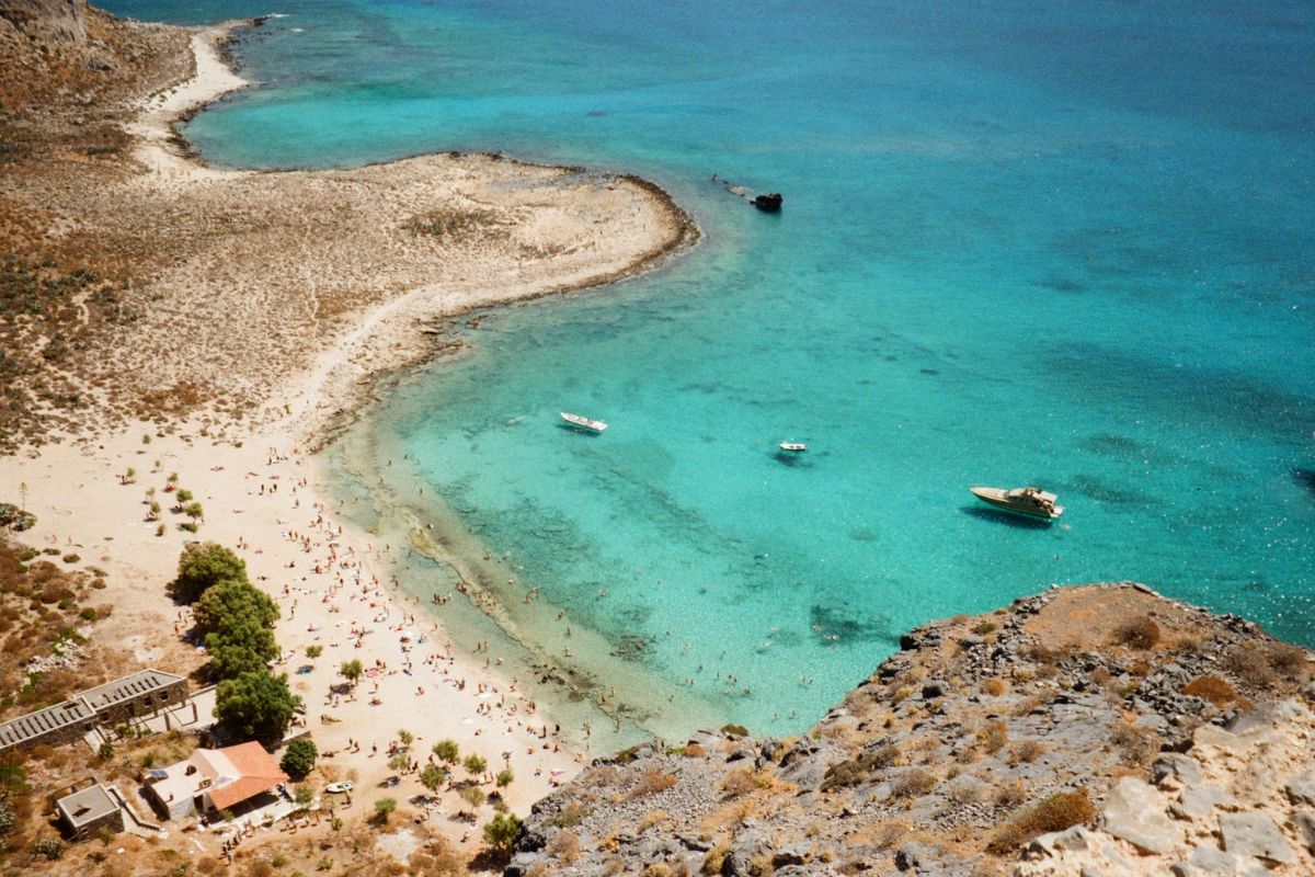 Aerial view of Balos Beach in Crete, with turquoise shallow water, a sandy shoreline, swimmers, and boats offshore.