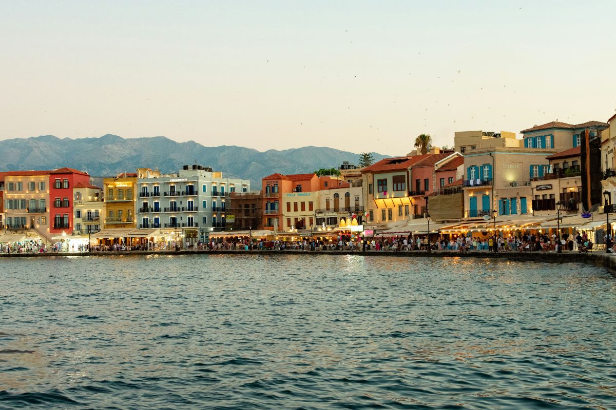 Chania Old Venetian Harbor in Crete at dusk, with colorful waterfront buildings, restaurants, and the White Mountains in the background.